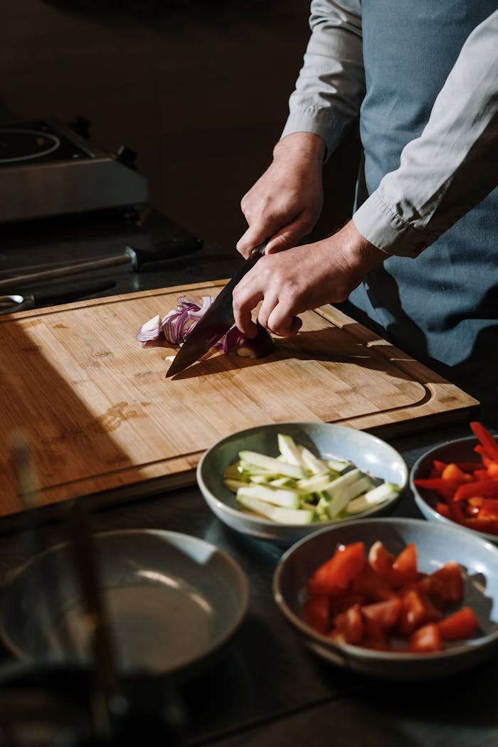 A chef skillfully slices onions on a wooden board with fresh vegetables ready in bowls.